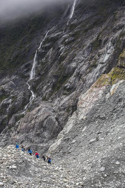 Hikers approaching the face of Franz Josef Glacier are dwarfed by the cliffs of "schist" rock.